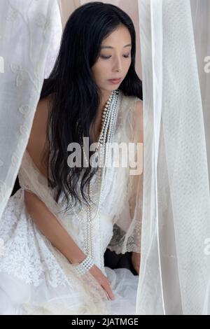 Une belle femme asiatique portant une robe en dentelle blanche et posant parmi le tissu transparent coulant Banque D'Images