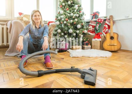 Une jeune femme qui nettoie avec un aspirateur, passe l'aspirateur sous les aiguilles de Noël avec des ornements du nouvel an sur un parquet en bois dur. Banque D'Images