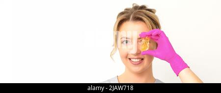 Le maître de l'enlèvement de cheveux de sucre tient la pâte de sucre jaune liquide, couvrant son oeil avec de la cire pour l'épilation sur un fond blanc Banque D'Images