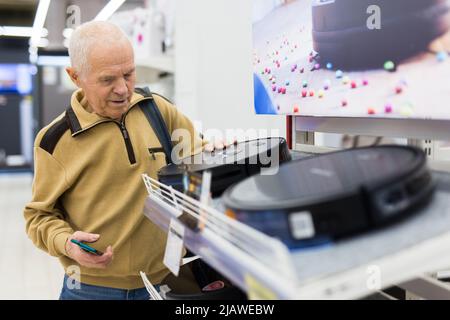 Sénior homme retraité achetant un aspirateur robotique dans la salle d'exposition du magasin d'appareils électriques Banque D'Images