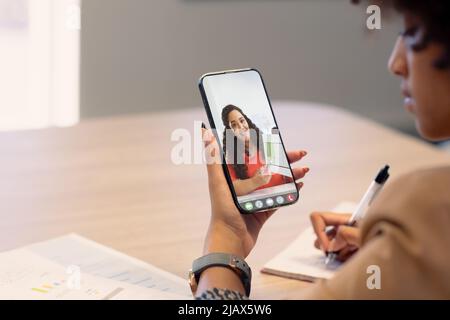 Femme d'affaires biraciale prenant des notes pendant un appel vidéo avec une collègue biraciale féminine montrant des données Banque D'Images