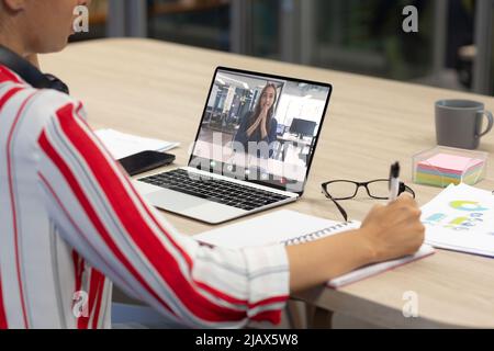 Femme d'affaires caucasienne écrivant sur un agenda lors d'une réunion en ligne avec une collègue caucasienne Banque D'Images