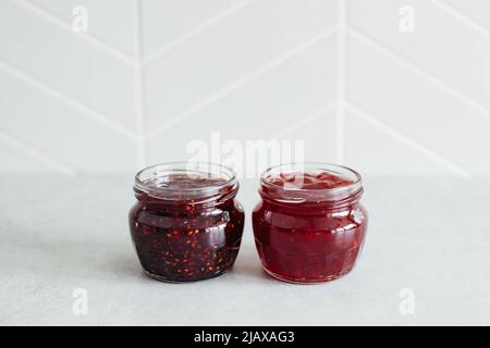 Deux pots ouverts en verre avec confiture de fraises rouges et de framboises sur table grise contre mur de cuisine en carreaux gris. Vue avant, espace de copie, mise au point sélective. Banque D'Images