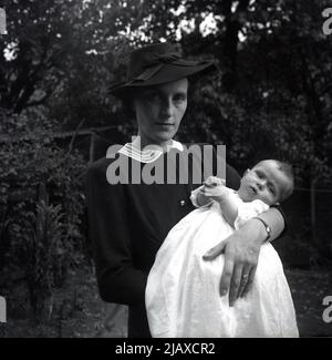 1950s, historique, debout dans une époque de jardin, une femme élégamment habillée, portant un chapeau, tenant son bébé à son baptême, Angleterre, Royaume-Uni. Banque D'Images