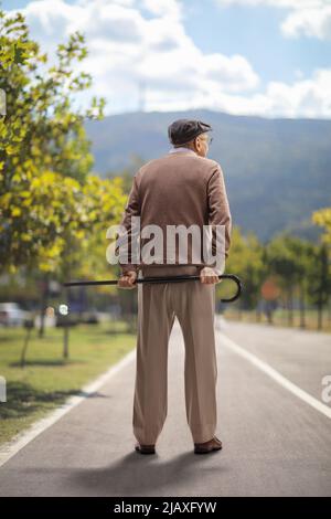 Vue arrière sur toute la longueur photo d'un homme âgé debout et tenant une canne derrière son dos à l'extérieur Banque D'Images