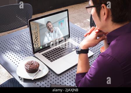 Jeune homme asiatique assis avec la main sur le menton tout en parlant à un médecin de race blanche mature sur ordinateur portable Banque D'Images