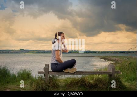 Une seule femme assise sur un banc à côté de l'eau et écoutant de la musique sur son casque. Banque D'Images