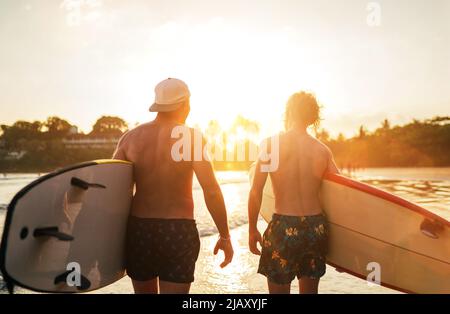 Père avec un fils adolescent marchant avec des planches de surf près de la plage de sable de l'océan avec des palmiers sur fond éclairé par le soleil de coucher de soleil.Ils sourient et ha Banque D'Images