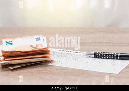 Sur une table en bois se trouve un billet de loterie sur lequel sont empilés plusieurs billets de 50 euros et un stylo. Banque D'Images