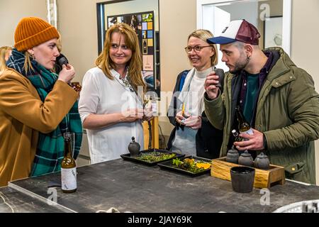 Plusieurs personnes dégustent des herbes et des boissons ensemble à une table ici dans le magasin SÓLEY à Reykjavik, Islande : une femme avec un bonnet orange, une femme (peut-être Sóley Elíasdóttir, fondatrice de SÓLEY), une femme portant des lunettes et un homme avec une casquette de baseball. De petits bols de herbes séchées, des tranches d'orange et plusieurs bouteilles sont posés sur la table. Banque D'Images