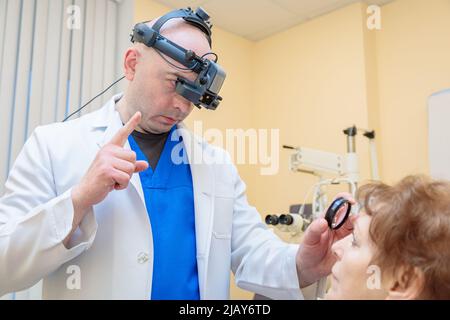 Un ophtalmologiste mâle vérifie la vue d'une femme adulte avec un ophtalmoscope binoculaire. Banque D'Images