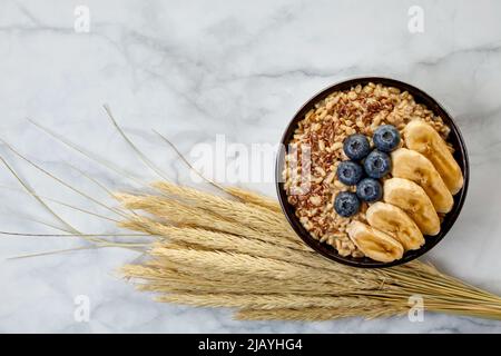 Flocons d'avoine entière avec bleuets et tranches de banane dans un bol sombre, entouré de grains saupoudrés, épis de maïs. Journée internationale du porridge Banque D'Images