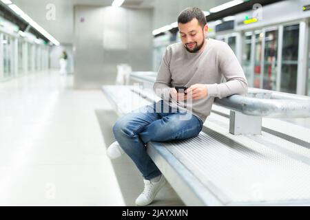 Homme barbu assis avec le téléphone sur le banc sur la plate-forme de métro Banque D'Images