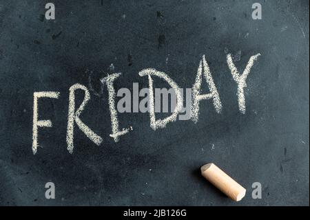 Vendredi. Nom du jour de la semaine écrit en craie jaune sur tableau noir. Texte manuscrit. Un morceau de craie colorée se trouve à côté. Banque D'Images