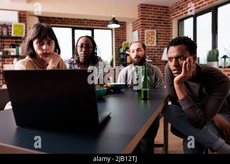 Des amis multiethniques anxieux assis à la maison tout en regardant des films de suspense ensemble sur un ordinateur portable. Les personnes dans le salon profitent d'activités de loisirs amusantes tout en prenant des en-cas et des boissons. Banque D'Images