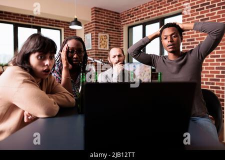 Divers groupes d'amis ont été étonnés de réagir aux événements tragiques qui se sont produits sur un ordinateur portable à la maison. Des gens multiraciaux inquiets assis à table dans le salon tout en regardant des films de suspense. Banque D'Images