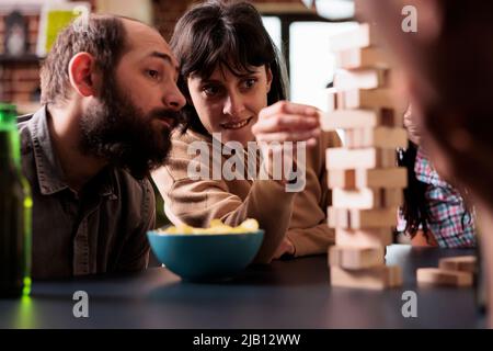 Des gens concentrés assis à une table dans le salon tout en jouant avec des blocs de bois. Femme concentrée qualifiée retirer soigneusement le cube en bois de la structure de la tour tout en appréciant les loisirs d'activité. Banque D'Images