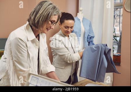 Portrait d'un designer sénior flou et d'une couturière hispanique d'âge moyen dans une veste élégante tenant un tissu bleu pour créer une nouvelle collection de Banque D'Images