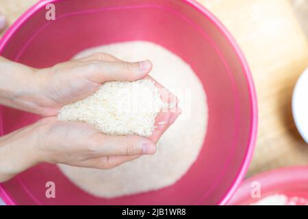 Vue de dessus une femme tenant le riz à la main au-dessus du bol en plastique rouge. Crise alimentaire mondiale. Riz sec cru. Riz blanc moulu non cuit. Zakat et concept de charité. Banque D'Images