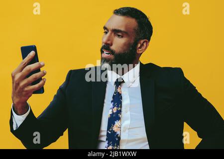 portrait de l'homme barbu expressif en colère en costume et cravate avec smartphone vidéo appel studio jaune d'arrière-plan Banque D'Images