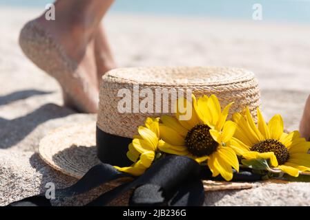 Jambes floues d'une femme marchant sur le sable de la plage en arrière-plan avec un chapeau de paille et des fleurs en gros plan. Concept de repos d'été. Banque D'Images
