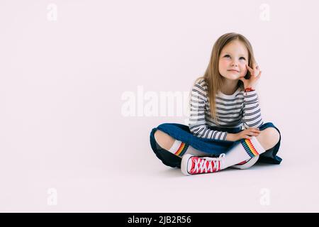 Indifférent sérieux cute blonde petite fille assise sur le sol, les jambes croisées, l'air ennuyé et unamused, ennuyeux, mal du pays ne veut pas faire ses devoirs, posent white Banque D'Images