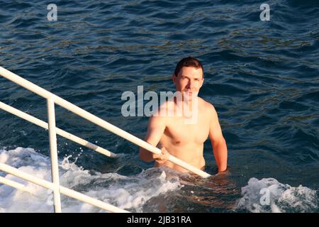 Vacances à la plage, baignade dans la mer. Portrait d'un beau homme musclé montant sur une échelle à partir des vagues bleues Banque D'Images