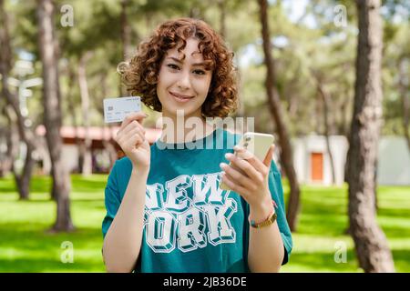 Jeune femme à tête rouge portant un t-shirt vert sur le parc de la ville, à l'extérieur montrant une carte de crédit ou de débit en plastique tout en tenant un téléphone portable. Boutique en ligne Banque D'Images