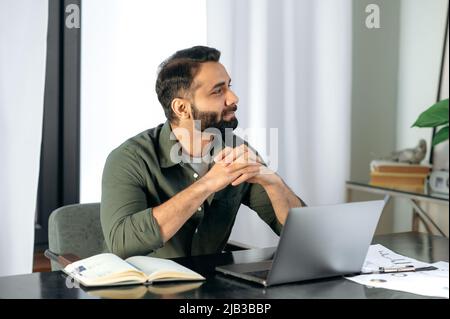 Un homme indien ou arabe bien pensé et réussi en tenue décontractée, courtier, gestionnaire, assis à un bureau dans le bureau, regarde pensivement loin, planifie une stratégie financière, évalue les risques, pense au projet Banque D'Images