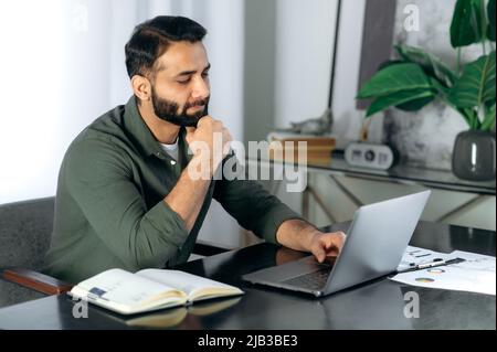 Conçu un homme indien ou arabe sérieux portant des vêtements décontractés, employé de bureau ou indépendant, assis à l'ordinateur de bureau dans le bureau moderne, regarde l'ordinateur portable attentivement, pense au projet Banque D'Images