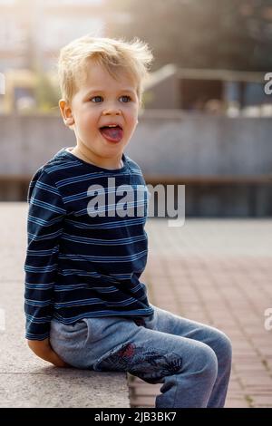 Un portrait d'un petit garçon méchant et sympathique assis près d'une fontaine de la ville, l'enfant pose, montrant sa langue. Banque D'Images