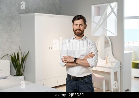 Vue de face d'un beau vendeur, concepteur, architecte debout avec des mains croisées dans la chambre. Brunette homme avec la barbe de concevoir, regarder l'appareil photo, sourire. Concept de mobilier. Banque D'Images