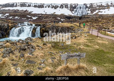 Un panneau indiquant « Bæjarfoss » se dresse devant la cascade du même nom, tandis que le grand Dynjandi (également appelé Fjallfoss) dévale les terrasses rocheuses en arrière-plan, Vestfjords, Islande. Bæjarfoss est l’une des six cascades de la cascade de Dynjandi à Arnarfjörður, protégée en tant que monument naturel depuis 1981. Les visiteurs marchent le long d’une promenade en bois entre les chutes. Banque D'Images