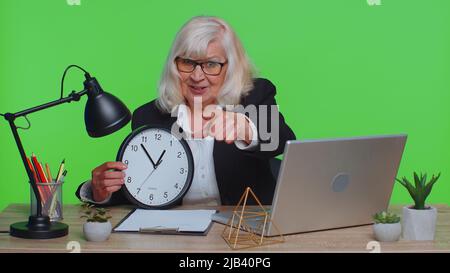 C'est votre temps. Femme d'affaires senior montrant l'heure sur l'horloge murale du bureau, ok, pouce vers le haut, approuver, pointer le doigt vers la caméra. Une femme âgée s'assoit au bureau isolé à l'intérieur, sur un studio de clé de couleur verte Banque D'Images