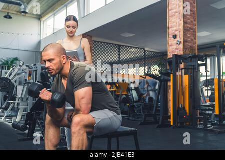 Homme millenial pensif fatigué dans un t-shirt vert et short gris clair assis et essayant de soulever une lourde haltères. Une femme européenne attirante debout derrière lui. Photo de haute qualité Banque D'Images