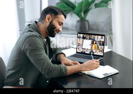 Vue latérale d'un homme intelligent qui a réussi à écouter une conférence en ligne, à prendre des notes dans un ordinateur portable, sur un écran d'ordinateur portable, un enseignant et un groupe de personnes multiraciales. Formation en ligne, webinaire Banque D'Images