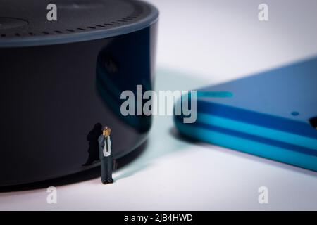 Homme d'affaires miniature debout devant un haut-parleur intelligent et appelant avec son téléphone. Un smartphone bleu se trouve à côté de l'appareil. Banque D'Images