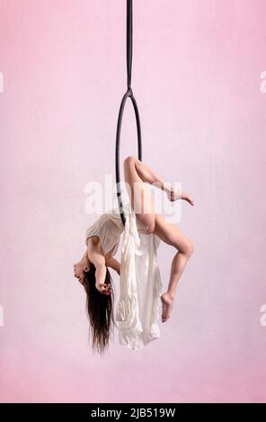 Vue latérale de la gymnaste femelle pieds nus en robe faisant un tour sur le cerceau aérien pendant la performance sur fond blanc et rose Banque D'Images