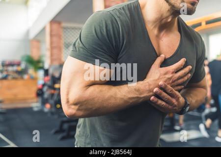 Tir sans tête dans une salle de gym d'un homme d'âge moyen caucasien barbu dans un t-shirt décontracté tenant ses mains sur sa poitrine, ayant une crise cardiaque. Photo de haute qualité Banque D'Images