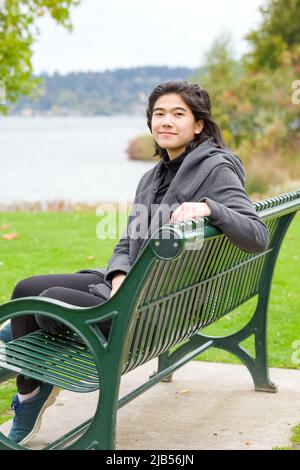 Jeune fille adolescente biraciale souriante ou jeune femme adulte en veste grise assise sur un banc de parc vert près du lac le jour couvert de l'automne ou du printemps Banque D'Images