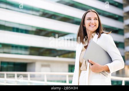 Femme d'affaires inspirée dans une tenue élégante et décontractée portant un ordinateur portable à pied dans la rue, souriante employée de bureau attirante regardant à l'extérieur, femme en costume avec ordinateur portable dans le centre-ville Banque D'Images