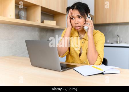 Une jeune femme agacée parle sur le smartphone en utilisant un ordinateur portable pour le travail à distance depuis la maison, une femme irritée regarde l'écran de l'ordinateur portable en ayant la conversation téléphonique Banque D'Images