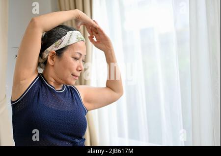 Portrait d'une femme asiatique d'âge moyen en bonne santé qui fait de l'exercice, fait du yoga, étire ses bras, s'entraîne à la maison. Banque D'Images