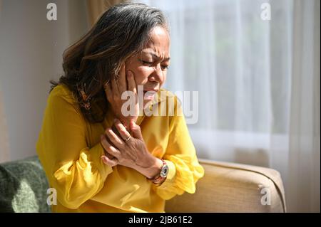 Une femme asiatique âgée touche des joues souffrant de douleurs dentaires soudaines. Vieille dame à expression douloureuse de dent. Concept de maladie dentaire Banque D'Images