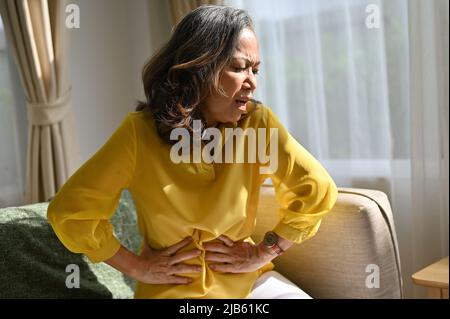 Malheureuse femme asiatique âgée sensation d'inconfort dans l'abdomen, maux d'estomac, gastrite pancréatite diarrhéas problème symptôme dans le salon. Banque D'Images