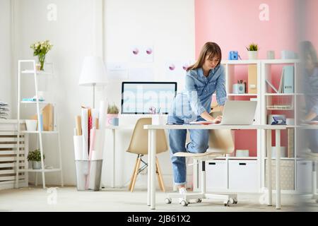 Contenu jeune femme dans des vêtements en denim debout à table au bureau et de vérifier les dossiers sur ordinateur portable Banque D'Images