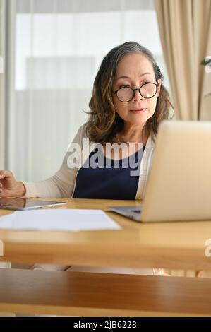 Portrait d'une femme d'âge asiatique ou d'une femme d'affaires attirante travaillant à distance dans sa maison, utilisant un ordinateur portable et examinant le rapport d'affaires. Banque D'Images