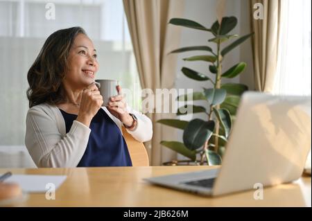 Souriante et élégante, femme de 60s ans et d'âge moyen appréciant de se reposer sur un fauteuil dans un salon moderne le matin. Banque D'Images