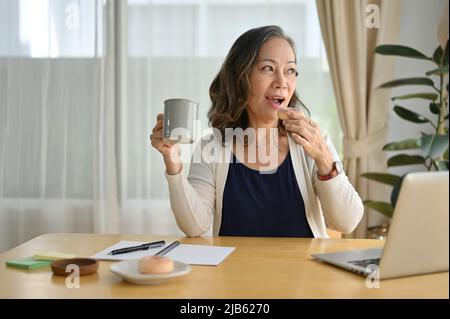 Une femme d'affaires asiatique âgée et joyeuse travaillant à distance à la maison, vous apprécierez de prendre un petit déjeuner avec café et beignets le matin. Banque D'Images