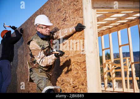 Les charpentiers martelent l'ongle dans le panneau OSB sur le mur du futur chalet. Hommes ouvriers construction maison en bois. Conception de la menuiserie et de la construction. Banque D'Images